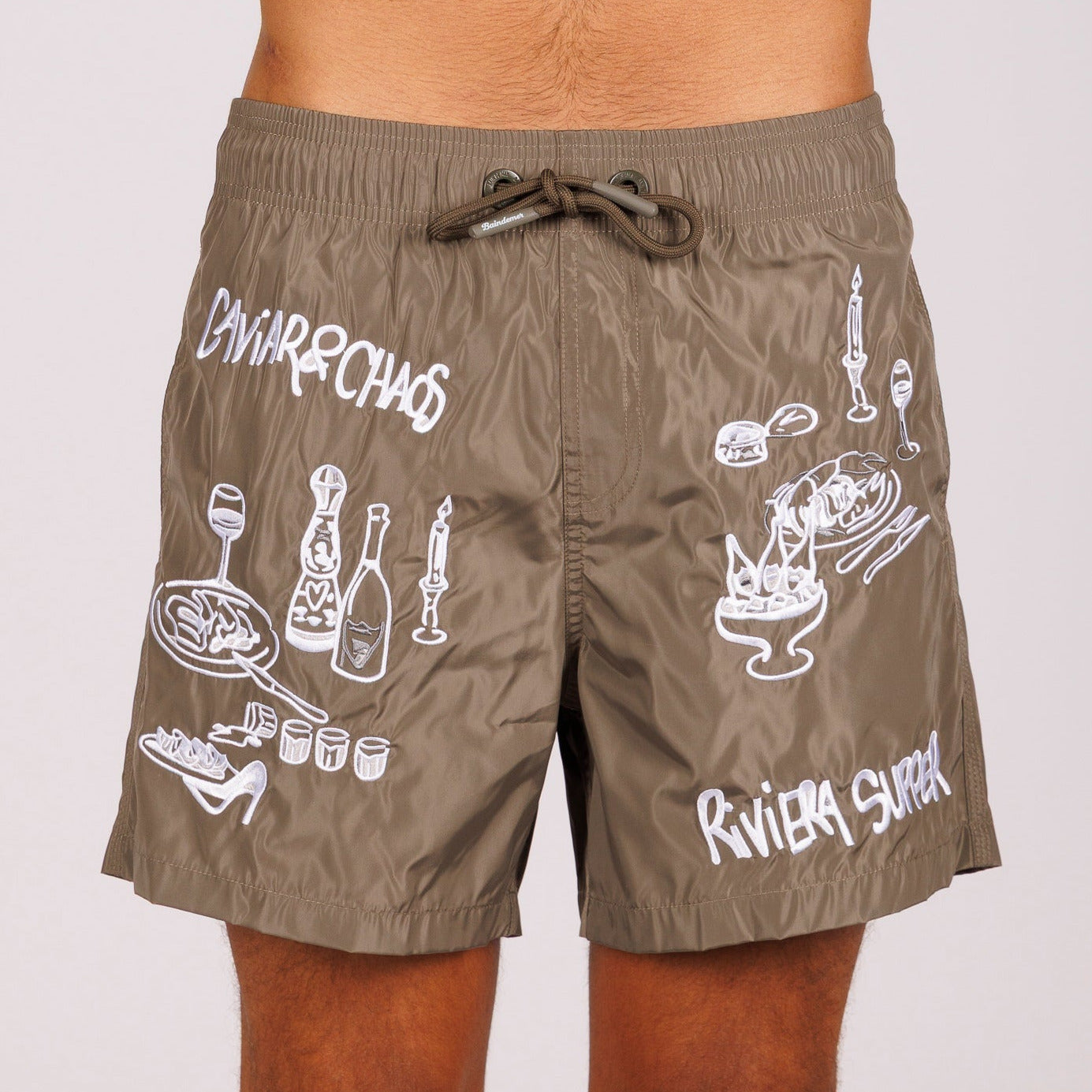Military green swim shorts front view with illustrated graphic and “Caviar&Chaos Riviera supper” text