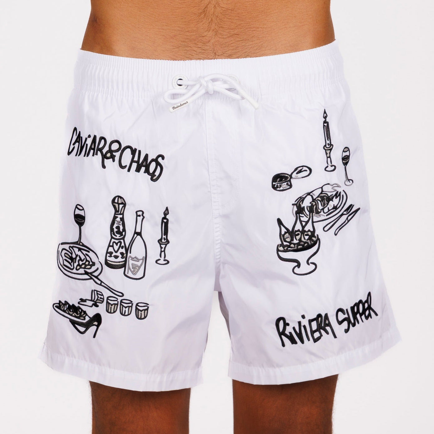 White swim shorts front view with illustrated graphic and “Caviar&Chaos Riviera supper” text