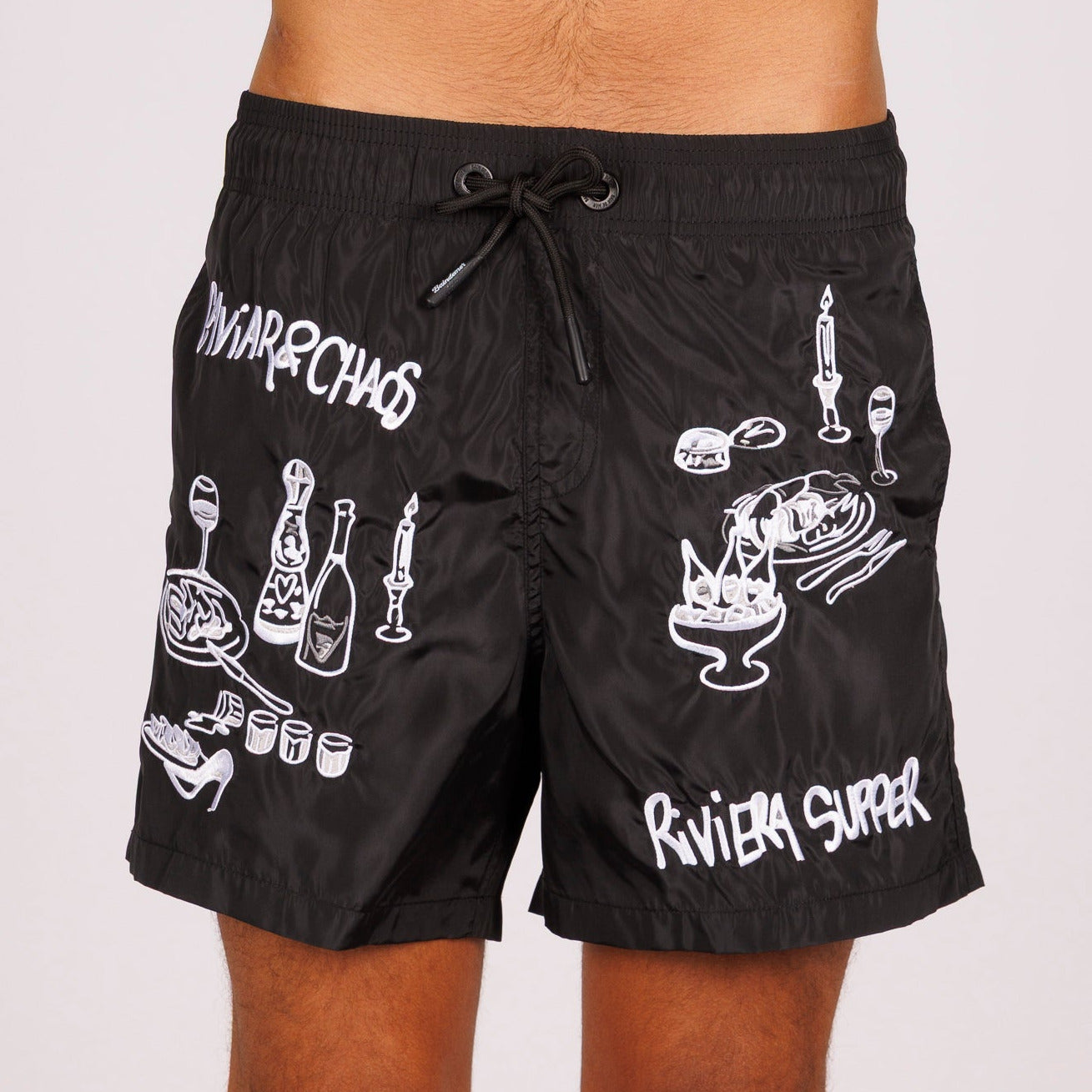 Black swim shorts front view with illustrated graphic and “Caviar&Chaos Riviera supper” text